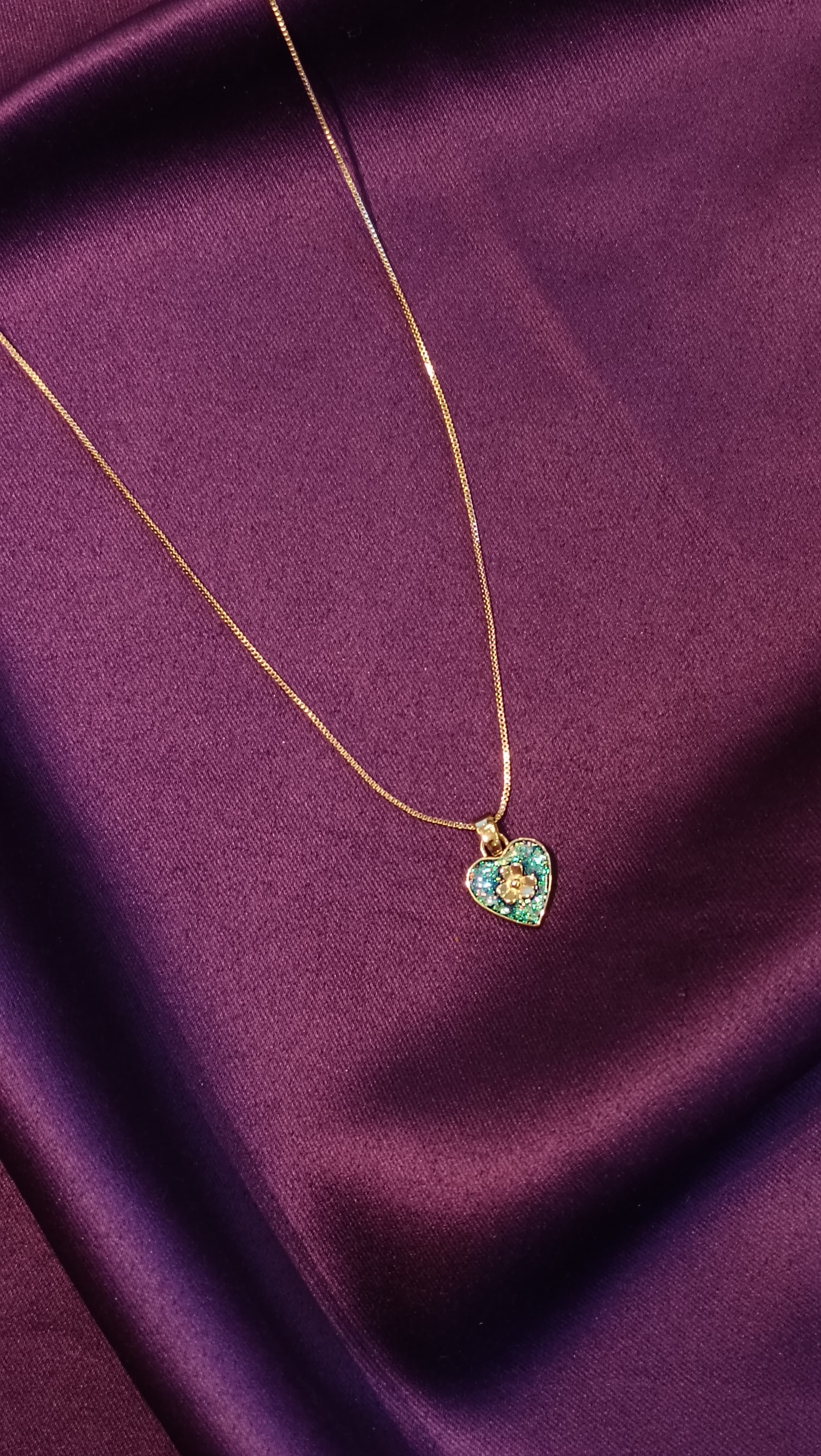 Gold Emerald Pavé Heart Pendant Box Chain Necklace close-up flat lay on purple fabric showing heart pendant detail and gold floral centre