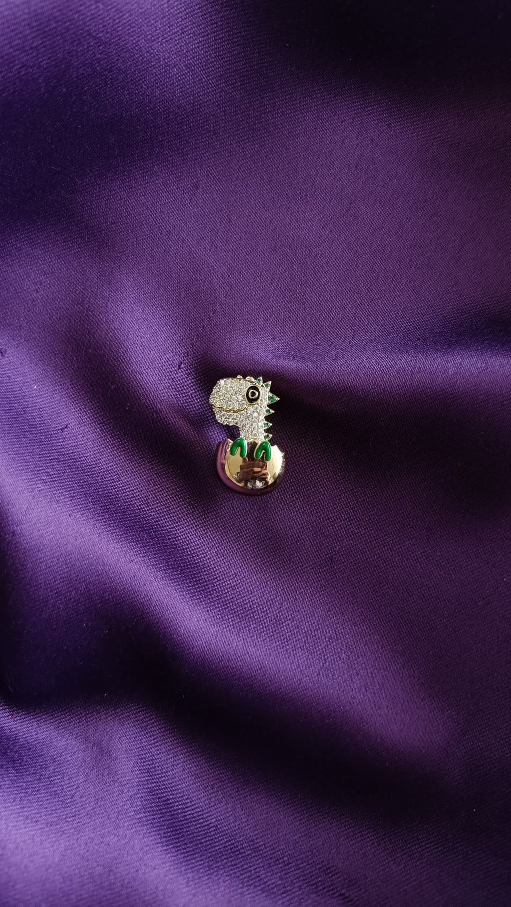 Gold Pavé Baby Dinosaur Hatching from Egg Lapel Brooch Pin front view flat lay on purple fabric showing white pavé dino head, green enamel spikes and gold egg