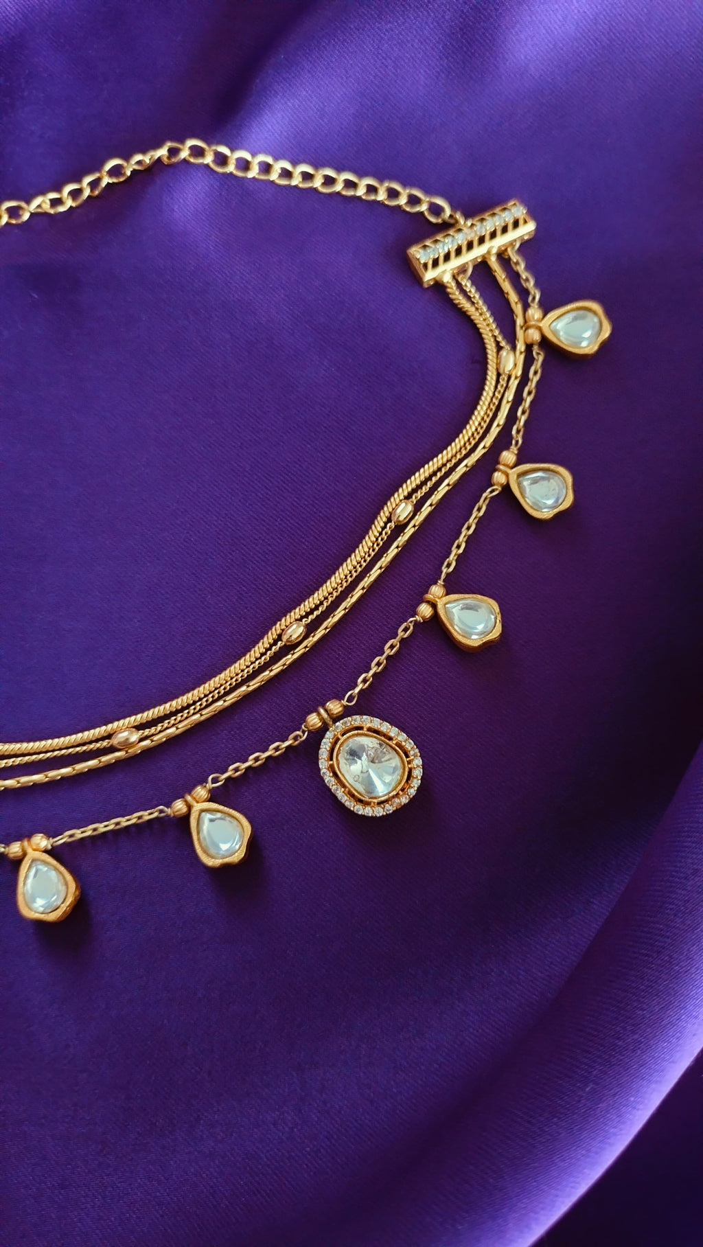 Gold Triple-Strand Polki Pear-Drop Charm Necklace close-up flat lay on purple fabric showing central pavé halo pendant and pear-drop charms